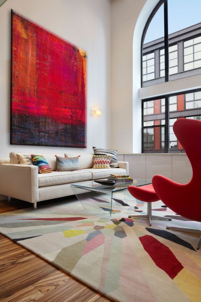 clean lined urban loft style interior with tall window, large abstract artwork, cream sofa, red egg chair and stool, and refracted colour on cream area carpet
