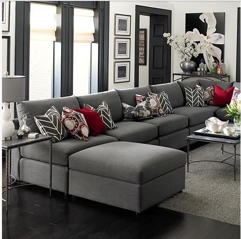 upscale living room with armless charcoal sectional sofa, area rug, red and grey cushions, Roman shades on the windows, and black trim moldings