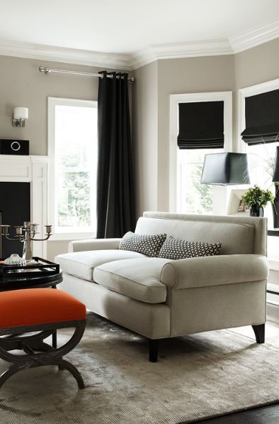 clean lined living room with ribbed neutral areal rug, sofa with single back cushion, orange ottoman, and black Roman shades and grommet style drapery