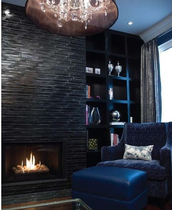 sitting area in upscale living room with fireplace with tile facing, built in black shelving, sheer drum chandelier, custom draperies, and blue armchair and ottoman