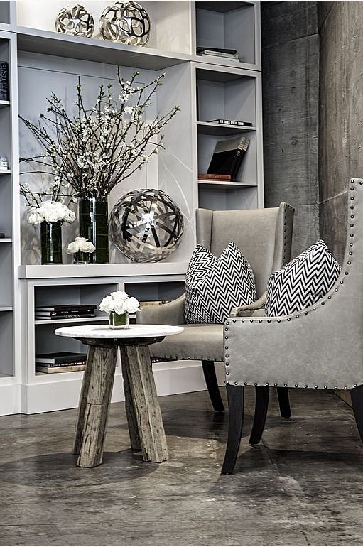 upscale loft style living room with concrete floors and walls, paired nail studded chairs in grey, reclaimed wood table, and wall unit in grey with silver accesories